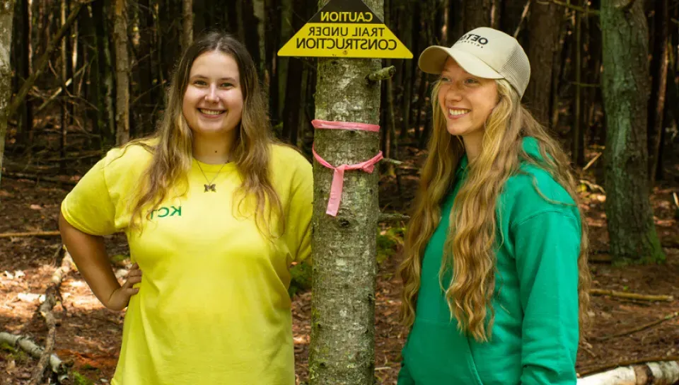 Two students standing in the woods next to a sign that reads, "Caution: Trail Under Construction"